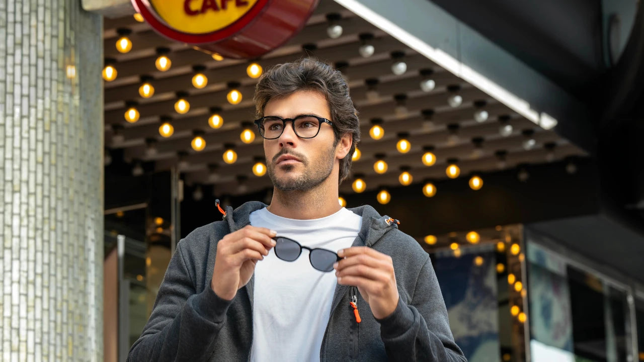 Man holding sunglasses in an urban setting with lights and a cafe sign.